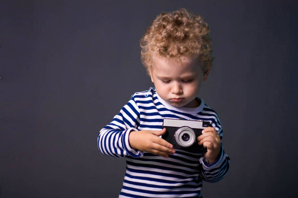 一个在灰色背景下有相机的小卷发男孩-一张摄影棚照片。孩子学会拍照.  图片素材