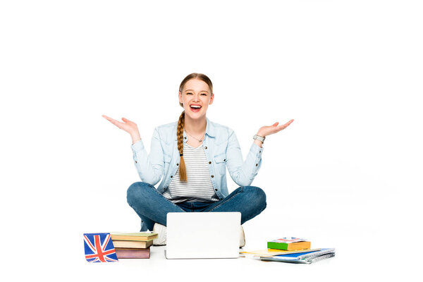 快乐的女孩坐在地板上，手里拿着笔记本电脑，书本和复印机，挂着英国国旗，在白色的墙上为自己做手势 图片素材