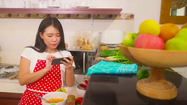 快乐的家庭烹饪-年轻快乐美丽的亚裔韩国家庭女厨师穿着红围裙，在手机里阅读健康食谱，在厨房里准备美味的饭菜 图片素材