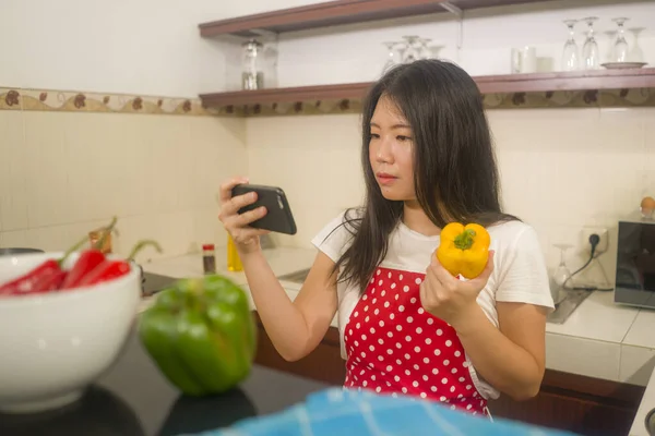 年轻、快乐、美丽的亚裔韩国家庭女厨师穿着红围裙阅读健康食谱，在手机上愉快地做饭，在厨房里准备美味的饭菜 图片素材