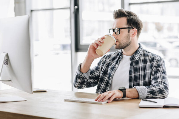 戴眼镜的年轻人喝咖啡从纸杯和使用台式计算机 图片素材