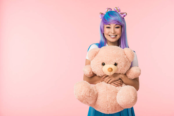 微笑的亚洲动漫女孩在假发抱着泰迪熊孤立在粉红色 图片素材