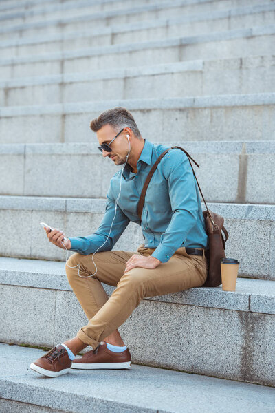 英俊的中年男子在太阳镜和耳机坐在楼梯上, 使用智能手机 图片素材