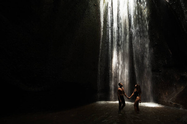 一对美丽的夫妇在一个山洞与瀑布。在瀑布下的溪流中，运动的男人和女人。一对性感的夫妇在热带瀑布下黄昏的光线。Tukad Chepung瀑布。复制空间 图片素材