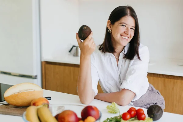 年轻快乐的女人手里拿着鳄梨，对着现代白色厨房里新鲜的水果和蔬菜笑着。健康食品概念。家庭烹饪 图片素材