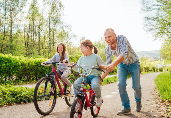 在户外散步的时候，带着两个女儿微笑的父亲。他教小女孩骑自行车.他们在夏季城市公园里聚精会神.快乐童年的概念形象. 图片素材